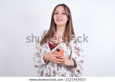 Smiling Young arab woman wearing floral shirt over white backgtound  friendly and happily holding mobile phone taking selfie in mirror.