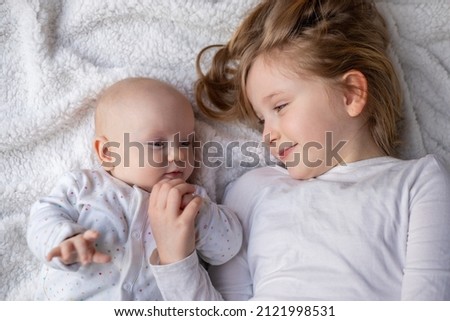 Happy children, toddler and older sister, hugging at home on a white blanket, smiling, shot from above. blonde girl hugging her newborn sister.