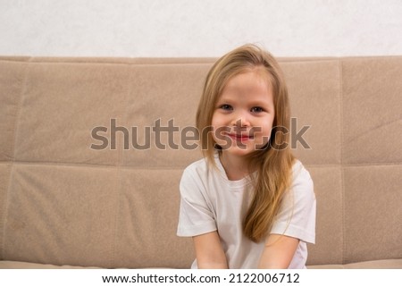 Portrait of a little blonde girl in a white T-shirt. Sitting on the couch and smiling
