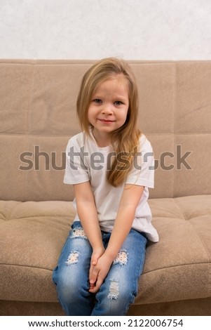 Portrait of a sad little blonde girl in a white T-shirt. Sitting on the couch unhappy