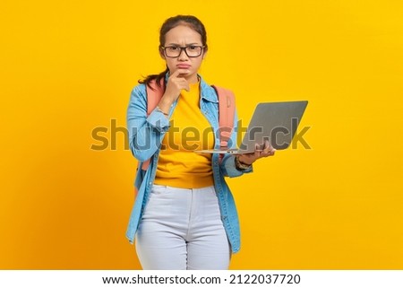 Portrait of serious young Asian woman student in casual clothes with backpack using laptop and thinking about question isolated on yellow background. Education in university college concept