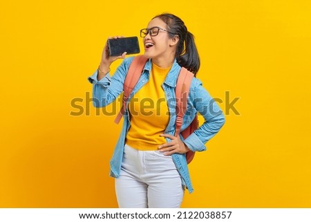 Portrait of smiling young Asian woman student in casual clothes with backpack enjoying singing using smartphone isolated on yellow background. Education in college university concept