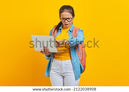 Portrait of surprised young Asian woman student in casual clothes with backpack using laptop and checking time on watch isolated on yellow background. Education in college university concept