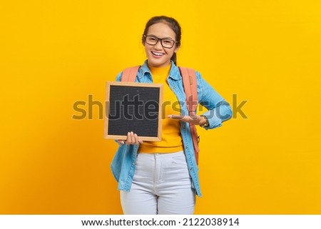 Portrait of smiling young Asian woman student in casual clothes with backpack pointing at blank chalkboard with palm isolated on yellow background. Education in college university concept