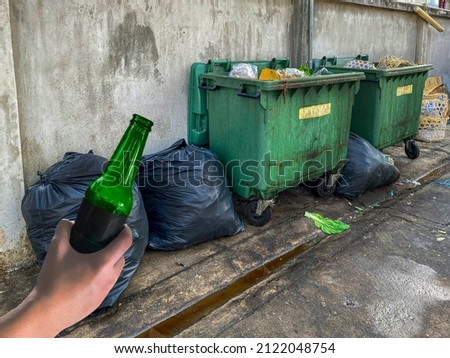 A man hold a green bottle to  dirty garbage containers and sewage on a residential street,  low technology development in the country,