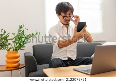 Middle age Asian man squint eye to look at the phone due to long eye sighted problems, which makes vision difficult.  Royalty-Free Stock Photo #2122078781