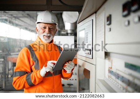 Portrait of professional factory worker or engineer in high visibility jacket and hardhat standing in control room of industrial production hall. Factory interior. Royalty-Free Stock Photo #2122082663