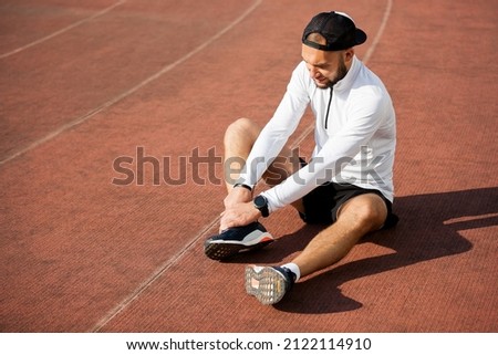 male runner in white black clothes holds his knee at the stadium, sports injury Royalty-Free Stock Photo #2122114910