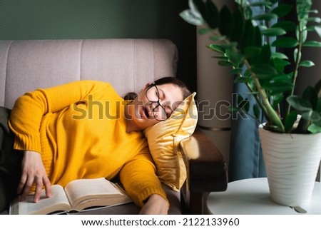 Middle-aged woman in glasses fell asleep while reading book at home, female sleeping with open mouth on sofa near coffee table with houseplant in living room. Sleep deprivation Royalty-Free Stock Photo #2122133960
