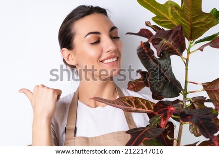 Young housewife in an apron on a white background holds a houseplant in a pot, smiles positively