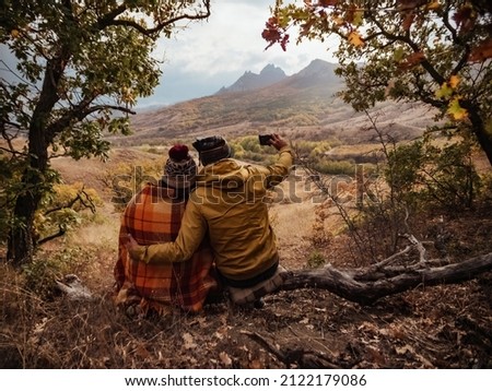 A man and a woman are resting in the mountains at a halt in the fall. The man takes a photo on the phone. View from the back.