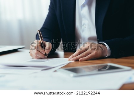 Close-up photo of businessman's hands signing documents at desk, man at work in business suit