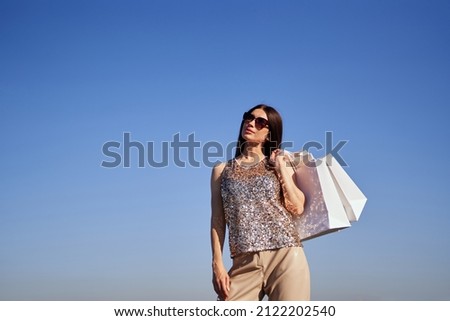 Happy caucasian woman with shopping bags with blue sky background. Attractive brunette glam girl in sunglasses with shopping bags standing in bright clothes at sunny day. High quality photo