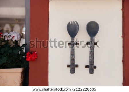 Large metal and old spoon and fork with carvings and patterns on them hang on the wall of cafe or restaurant on Santorini island, Oia, Greece. Signboard on the street near window and flower in pot