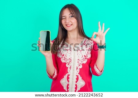 young beautiful moroccan woman wearing traditional caftan dress over green background holding in hands cell showing ok-sign
