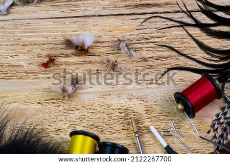 Several fly tying flies on a wooden background among fly tying materials. Fantasy dry flies for fishing. Royalty-Free Stock Photo #2122267700