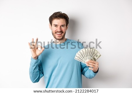 Handsome young man showing quick loans money, make okay gesture and smiling with cash, standing over white background