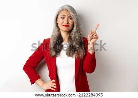 Disappointed and skeptical asian female entrepreneur pointing finger upper right corner, grimacing and frowning upset, showing bad promo, white background