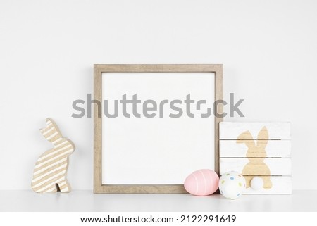 Mock up wood frame with Easter decor on a white shelf. Shabby chic wooden sign, egg, bunny. Square frame against a white wall. Copy space.