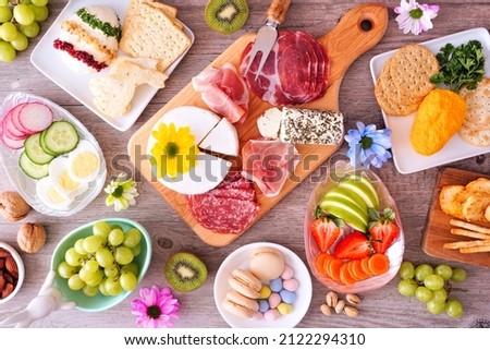 Spring or Easter theme charcuterie table scene against a wood background. Collection of cheese, meat, fruit and vegetable appetizers. Overhead view. Royalty-Free Stock Photo #2122294310