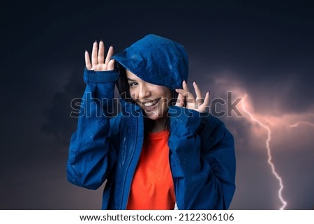 Portrait of a smiling girl dressed in blue raincoat in drops posing with hood against the background of a gloomy sky with lightning.