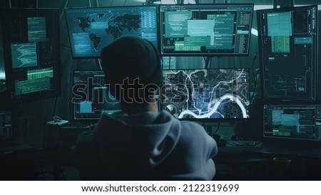 Teenage hacker with glasses watching a car on a monitor and pressing a keyboard in a dark cybercriminal hideout with lamps during a cyberattack Royalty-Free Stock Photo #2122319699