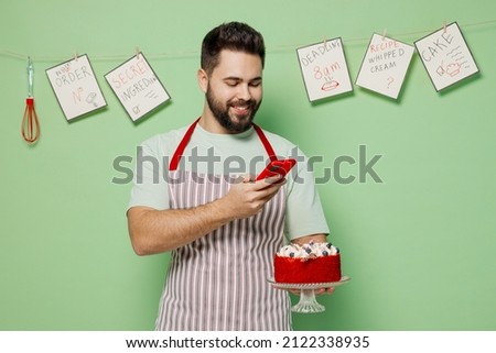 Young smiling happy male chef confectioner baker man in striped apron take photo of birthday sweet dessert cake isolated on plain pastel light green background studio portrait. Cooking food concept.