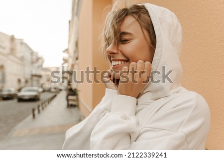 Pretty young caucasian woman in white hoodie smiles tenderly while standing on fresh sigh. Keeps eyes closed, feels comfortable and full. Self-love expression concept