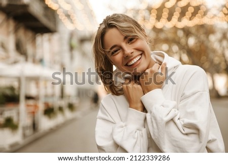 Close up portrait caucasian young happy woman with fresh and clean skin stands outside. Smiling blonde holds collar of white sweatshirt. Lifestyle, female beauty concept Royalty-Free Stock Photo #2122339286