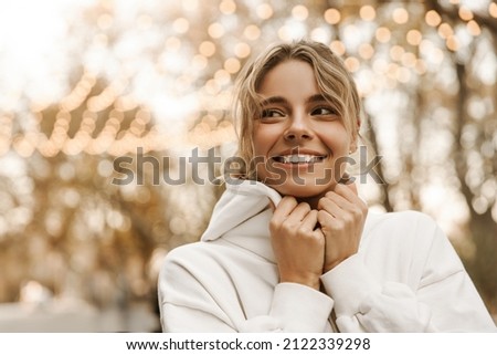 Close-up of pretty young caucasian woman in casual autumn clothes walking in city. Beautiful stylish lady is wrapped white sweatshirt, fashionable hat, autumn fashion trend. Street style concept. Royalty-Free Stock Photo #2122339298