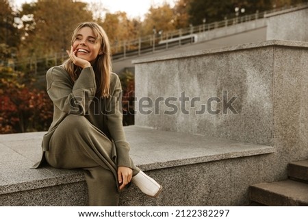 Portrait of tanned cheerful young woman is resting while sitting on concrete staircase in autumn day. Blonde girl with snow-white smile looks to side. People, season and leisure concept. Royalty-Free Stock Photo #2122382297