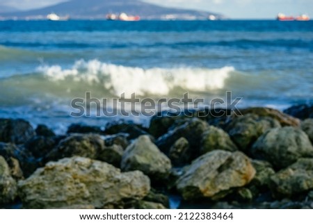 Blurred landscape, seashore with large stones, sea waves, white foam, blue water, sunlight, ships on the horizon line.