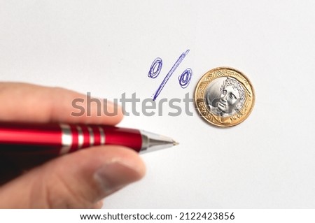 A Brazilian real coin worth 1 real and the percentage symbol to represent the interest rate and a man holding a pen. Concepts about Brazilian economy and investments.
