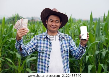 Handsome Asian male farmer holds Thai banknote money and smartphone at green maize field. Concept : Farmer gets agricultural supporting money via mobile app.                           Happy farmer.