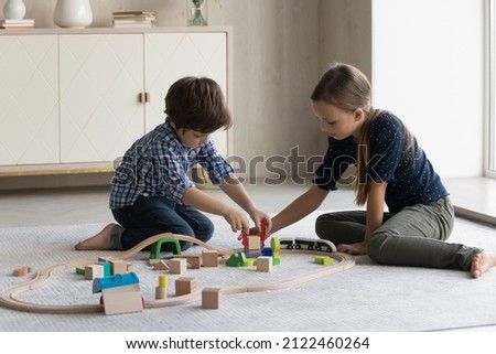 Two focused kids building toy railroad on floor, completing construction model. Little brother and sister sitting on heating floor at home, playing games with wooden blocks, training creative skills Royalty-Free Stock Photo #2122460264