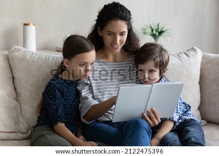 Happy engaged mom reading paper book to son and daughter. Mother and kids sitting on couch at home, enjoying leisure, children listening to exciting fairy tale. Family tome, literature, motherhood