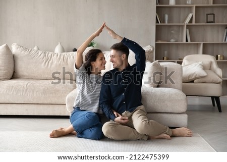 Happy homeowners celebrating relocation, moving into new home. Joyful married couple making hand roof shape above heads, sitting on floor in living room, smiling, laughing. House buying concept
