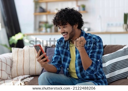 Overjoyed Indian man looking at the screen of mobile phone making yes gesture, feeling excited after receiving job offer, winning the lottery received a nice message in the mail, celebrates success Royalty-Free Stock Photo #2122488206