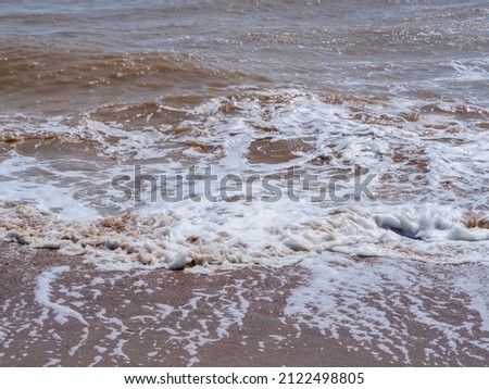 Brown muddy waves roll onto the shore with white foam, creating bizarre patterns.  Muddy surface of seawater due to strong wind waves and coastal erosion.  Abstract natural background. Royalty-Free Stock Photo #2122498805