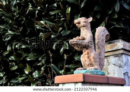Worn out statue of a squirrel on the top of a gate with a hedge as background
