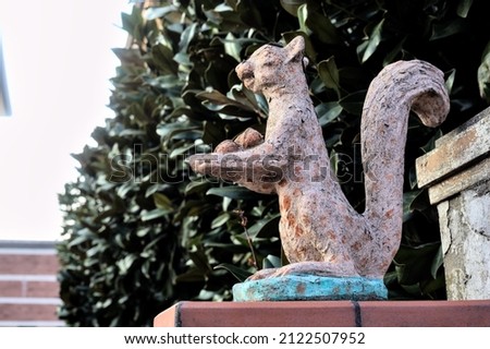 Worn out statue of a squirrel on the top of a gate with a hedge as background