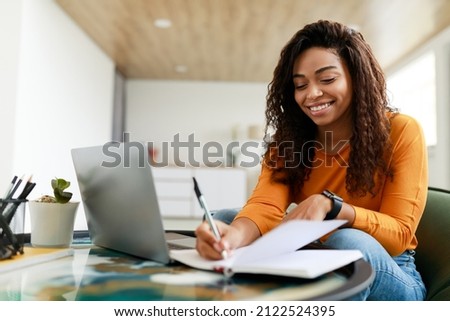 Business And Education Concept. Smiling young black woman sitting at desk working on laptop writing letter in paper notebook, free copy space. Happy millennial female studying using pc Royalty-Free Stock Photo #2122524395