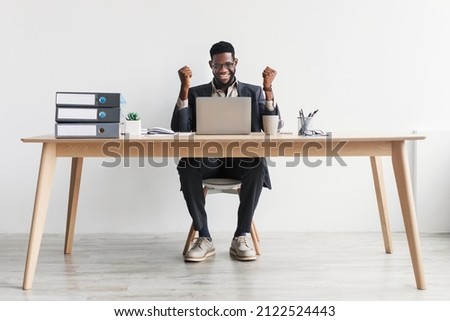 Full length of excited millennial African American office worker sitting at desk with laptop, making YES gesture against white studio wall. Young black businessman celebrating success at workplace Royalty-Free Stock Photo #2122524443