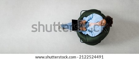People And Modern Technology. Above top view of young smiling black woman holding pc on lap sitting on bean bag in living room, looking up at camera. Lady browsing internet, free copy space panorama Royalty-Free Stock Photo #2122524491