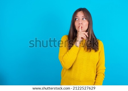 Young arab woman wearing yellow knitted sweater over blue backgtound silence gesture keeps index finger to lips makes hush sign. Asks not to share secret