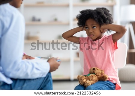 Unruly angry little black girl with bushy hair with teddy bear on her laps covering her ears and looking at psychologist unrecognizable woman, clinic interior. Child attending therapy session Royalty-Free Stock Photo #2122532456