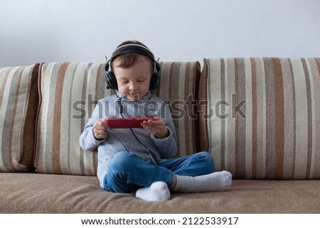 A 4-year-old boy in blue jeans and a gray turtleneck in headphones sits on the couch and looks enthusiastically at the phone