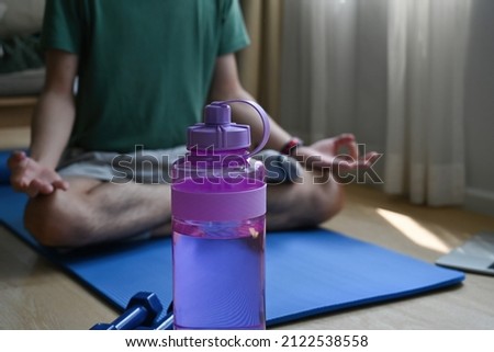 Close up focused water bottle in the foreground and a man sitting cross-legged meditating on a mat in a living room or yoga studio in the background, for home exercise , meditation and yoga concept.