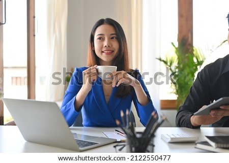 A frontal view portrait of a young smiley pretty Asian business woman looking away holding a cup of coffee working on a laptop in the office, for business and technology concept.