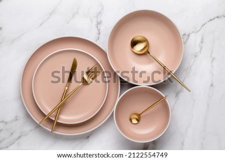Dishes and utensils for serving and eating meals. Beige round rimmed plates and gold colored cutlery on a white marble table, top view. Modern ceramic crockery, trendy tableware Royalty-Free Stock Photo #2122554749